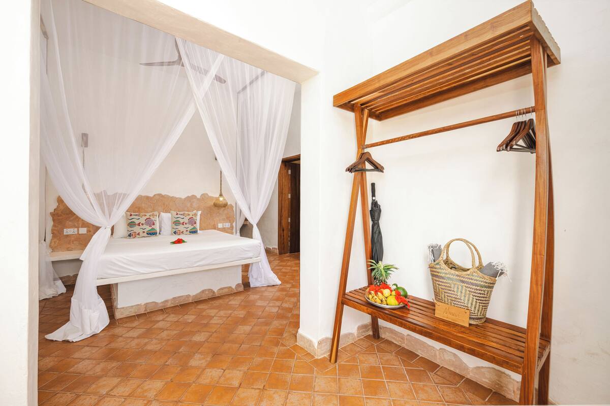 Une chambre à coucher avec un lit, un banc en bois surmonté d’un panier et un miroir.