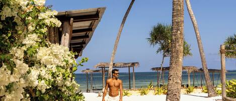 A beach scene with a person walking on the sand, palm trees, and a wooden structure.