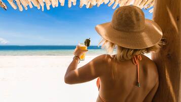 A person in a straw hat and bikini, holding a drink, under a wooden structure with a view of the beach and ocean.