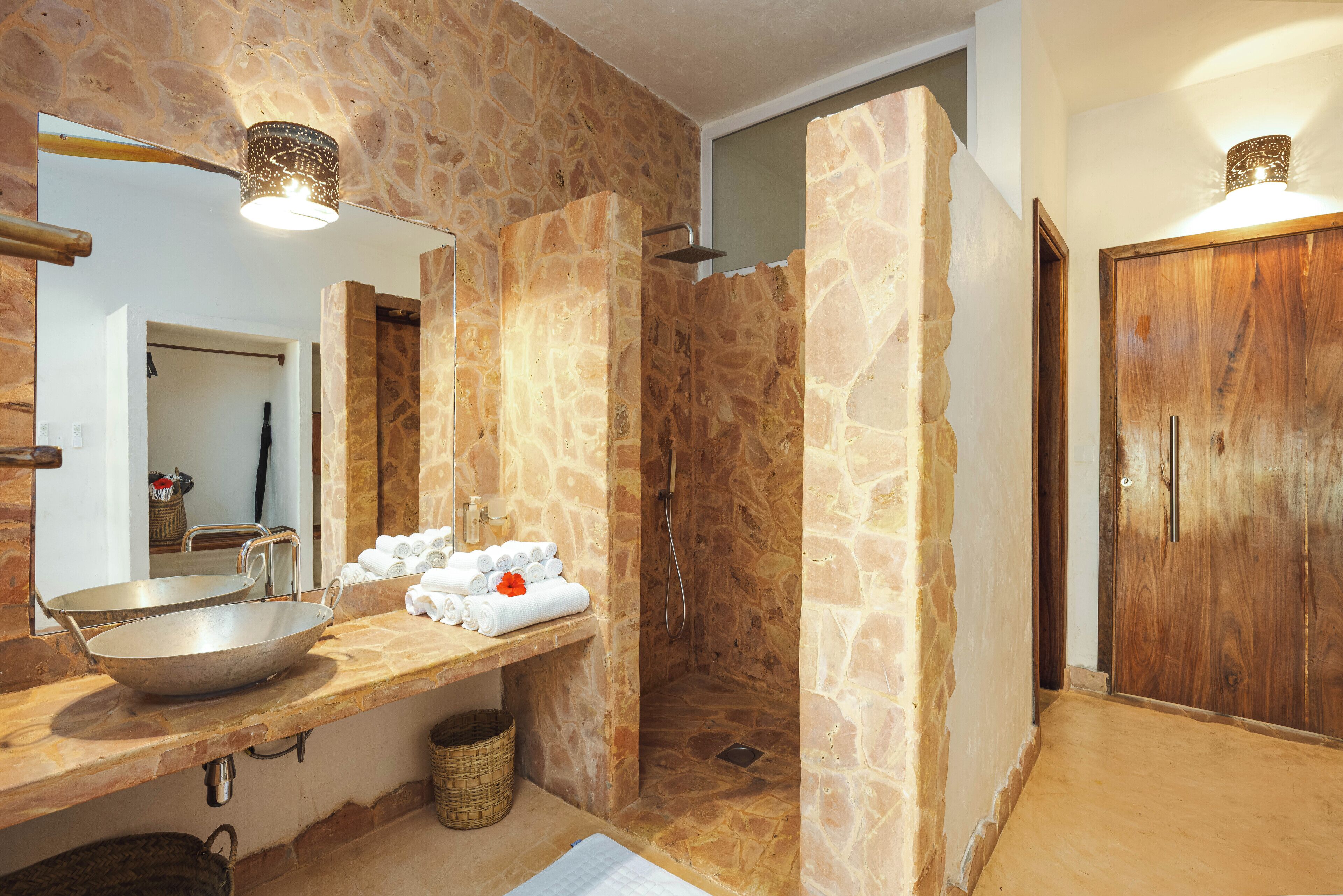 A bathroom with stone walls, a wooden door, a large mirror, a sink with a basin, and towels on a shelf.