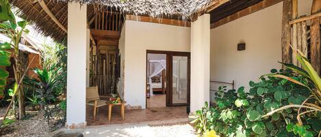 A thatched-roof outdoor area with a wooden bench, a small table with fruits, and a door leading to a room.