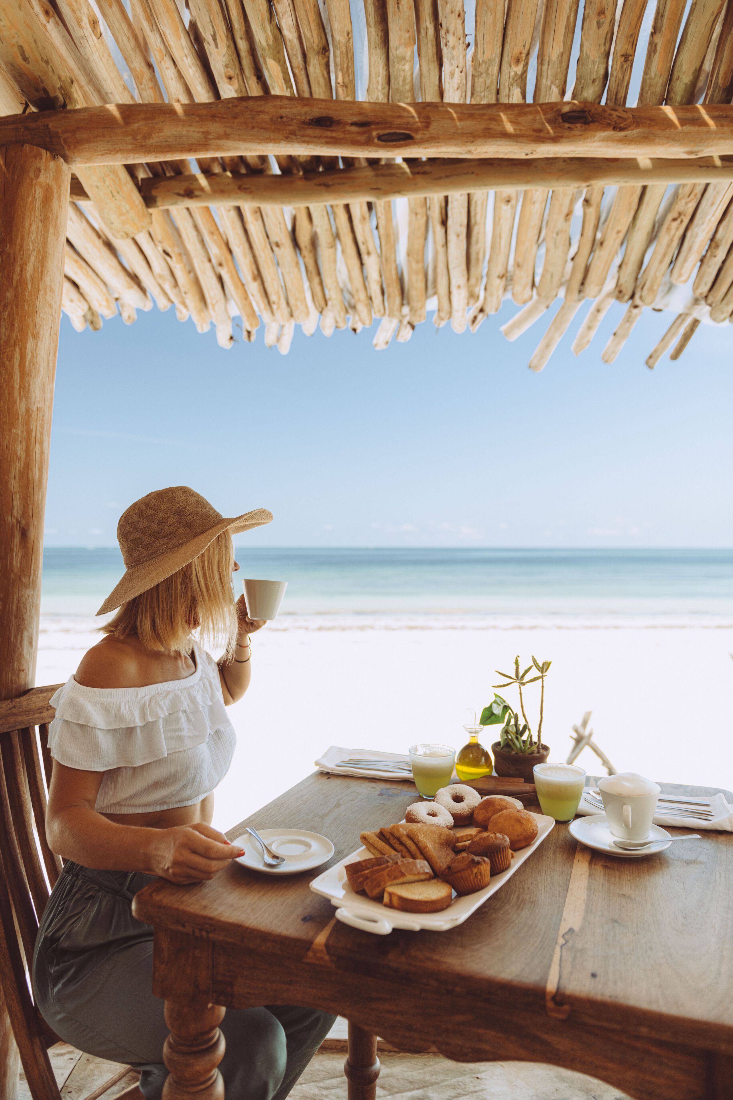 A person seated at a wooden table with a beach view, holding a cup, and a plate of pastries.