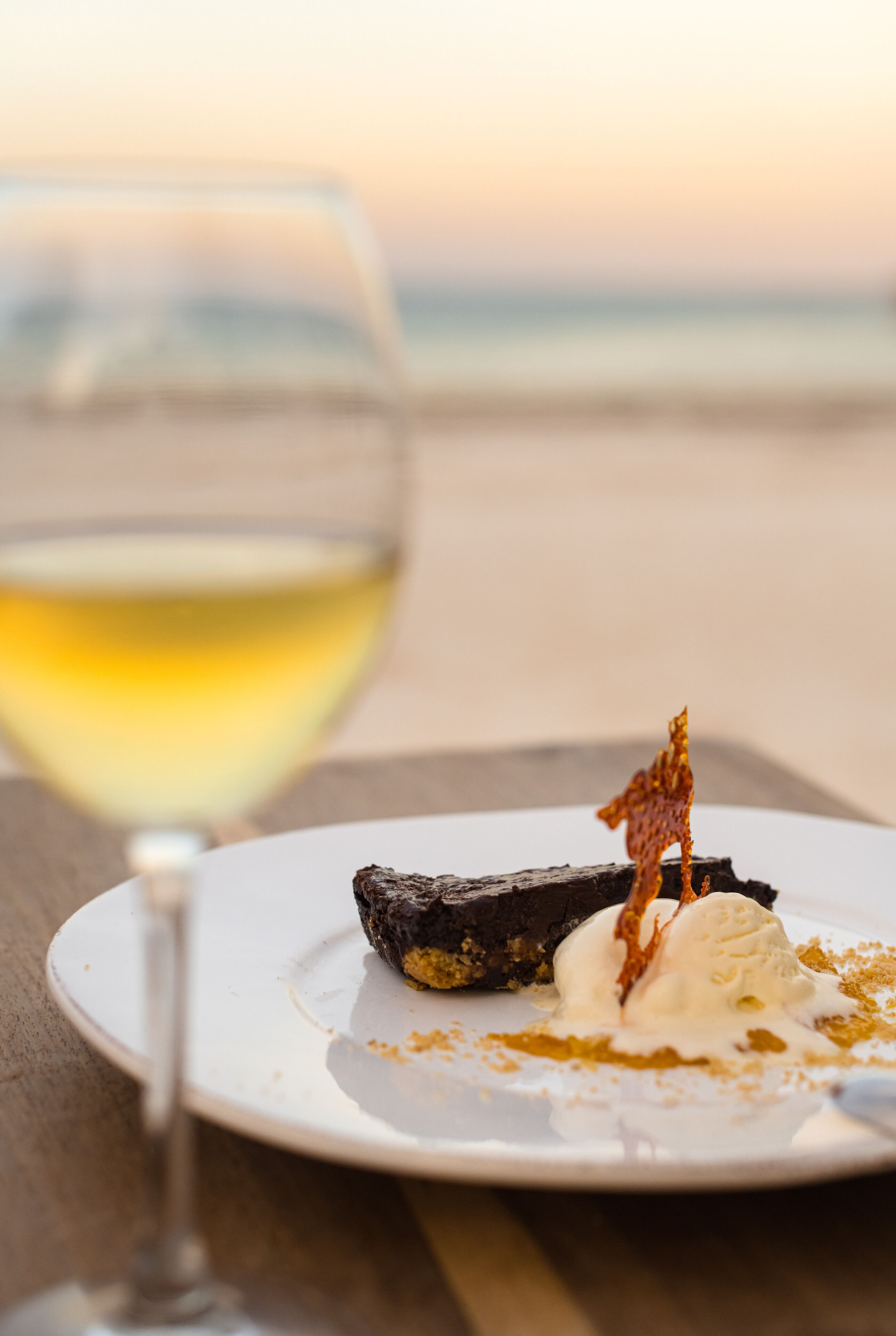 A dessert plate with a piece of cake and ice cream, accompanied by a glass of white wine, on a table with a blurred background.