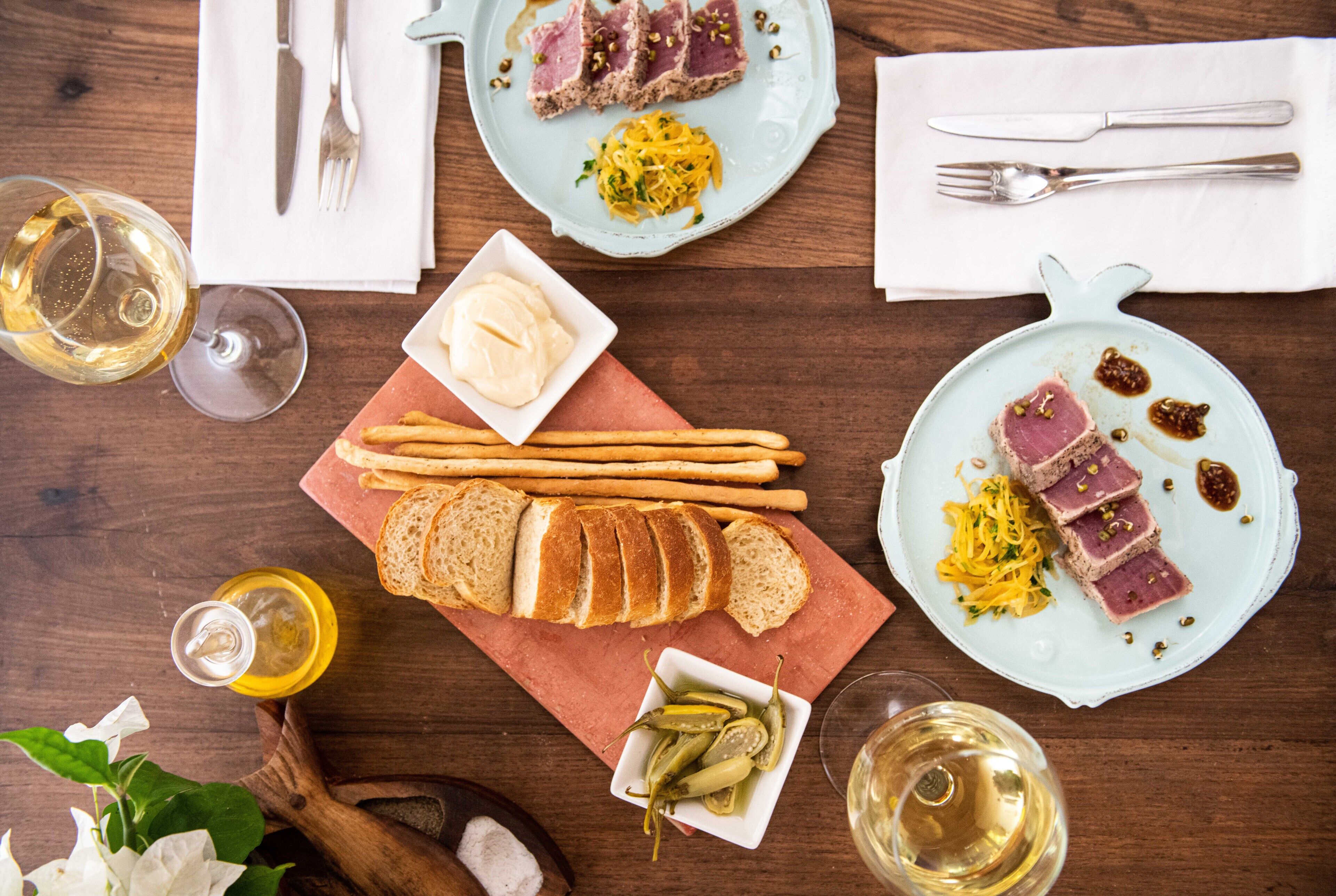 A wooden table with plates of food, including sliced meat, pasta, and bread, along with glasses of white wine.