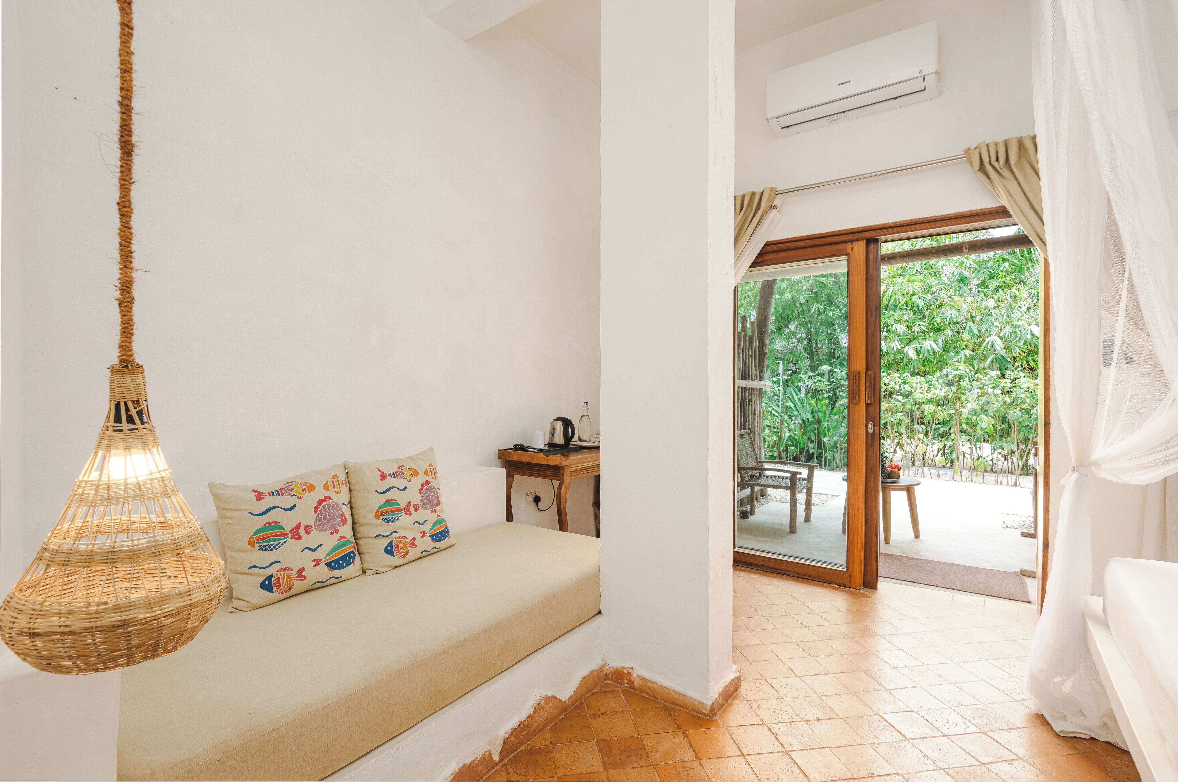 A room with a bed, a hanging light fixture, a small table with a kettle, and a view of a garden through a sliding door.
