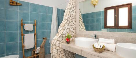 Un baño con cabina de ducha de piedra, toallero de madera, lavabo blanco y un espejo grande.