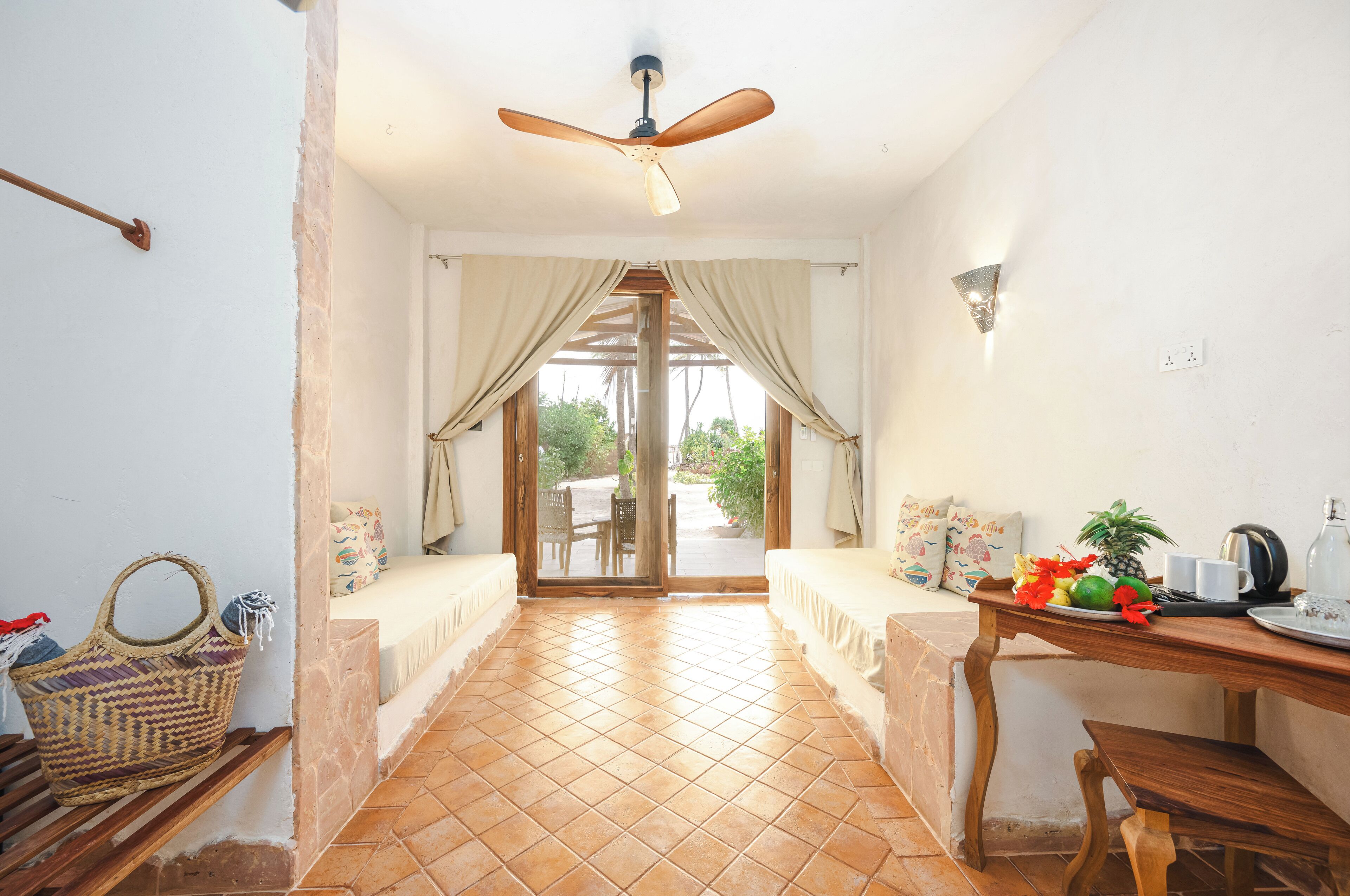 A room with a tiled floor, a ceiling fan, a wooden table with a tray of fruit, and a bench with cushions.