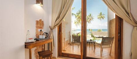 A room with a wooden desk, a chair, a bed, and a view of the beach through a sliding glass door.