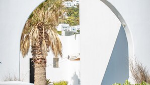 A white building with a stone wall and a palm tree in front. - Agia Irini (Santorini)