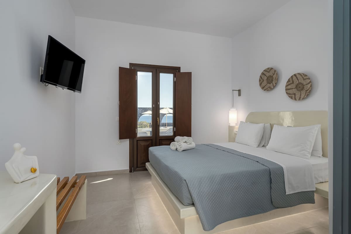 A hotel room with a bed, a TV mounted on the wall, a wooden bench, and a door leading to an outdoor area.