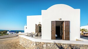 A white building with a stone foundation, a wooden door, and a view of the sea. - Agia Irini (Santorini)