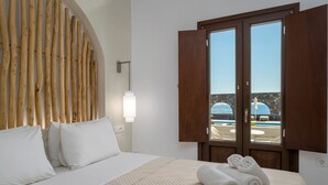 A hotel room with a bed, a wooden headboard, a window with a view of a pool, and a wall-mounted light fixture. - Agia Irini (Santorini)