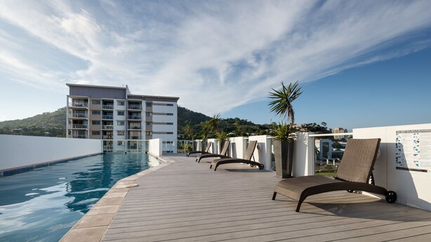 A poolside area with lounge chairs, a building in the background, and a clear sky. - Holborn at Central (Townsville)
