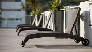 A row of sun loungers with wheels on a wooden deck, a white wall with a security camera, and a palm tree in the background. - Holborn at Central (Townsville)