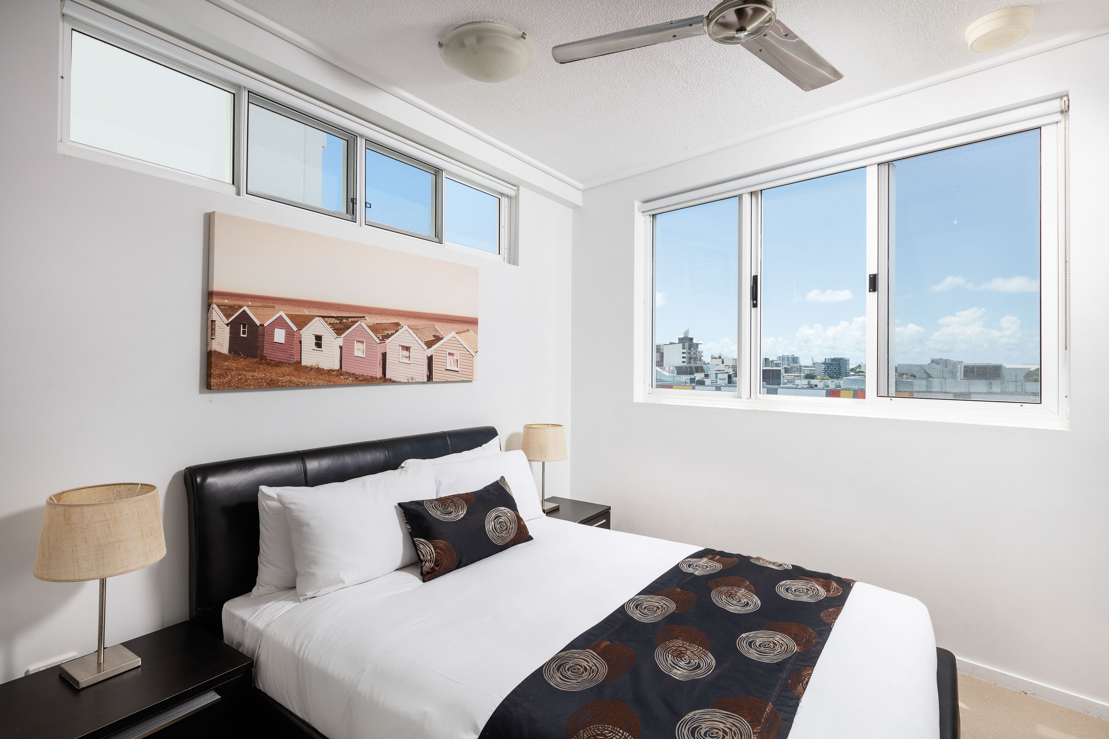 A modern hotel room with a large bed, a ceiling fan, a city view, and artwork on the wall.