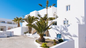 A white building with a curved entrance, palm trees, and a clear blue sky. - Nikos Hotel (Santorini)