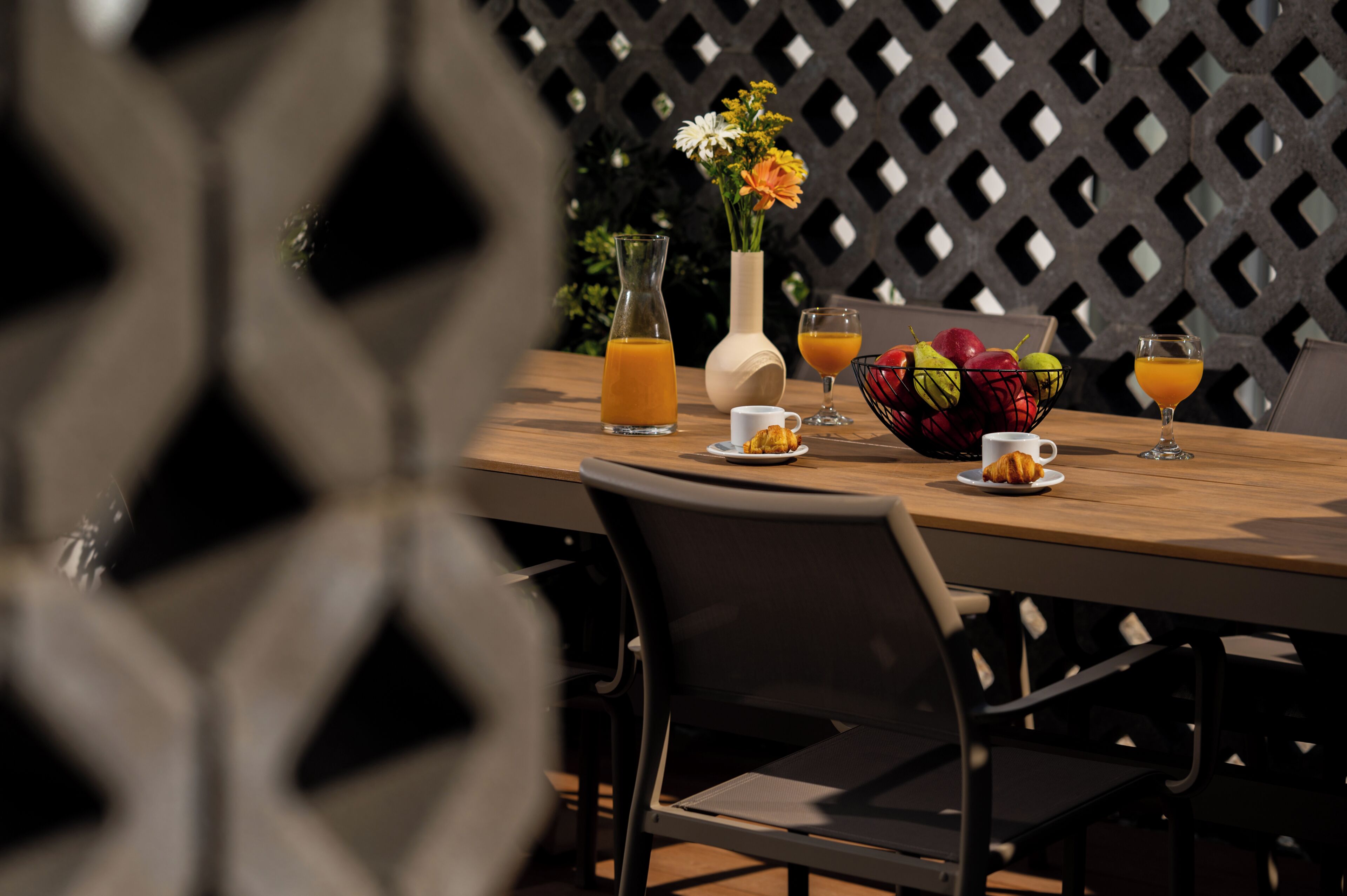 A wooden table with a glass pitcher, two glasses of orange juice, a white cup, a bowl of fruit, and a vase with flowers.