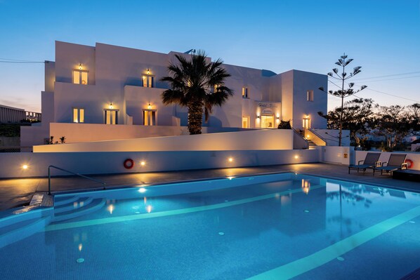 A modern building with a swimming pool, palm trees, and lounge chairs. - Nikos Hotel (Santorini)