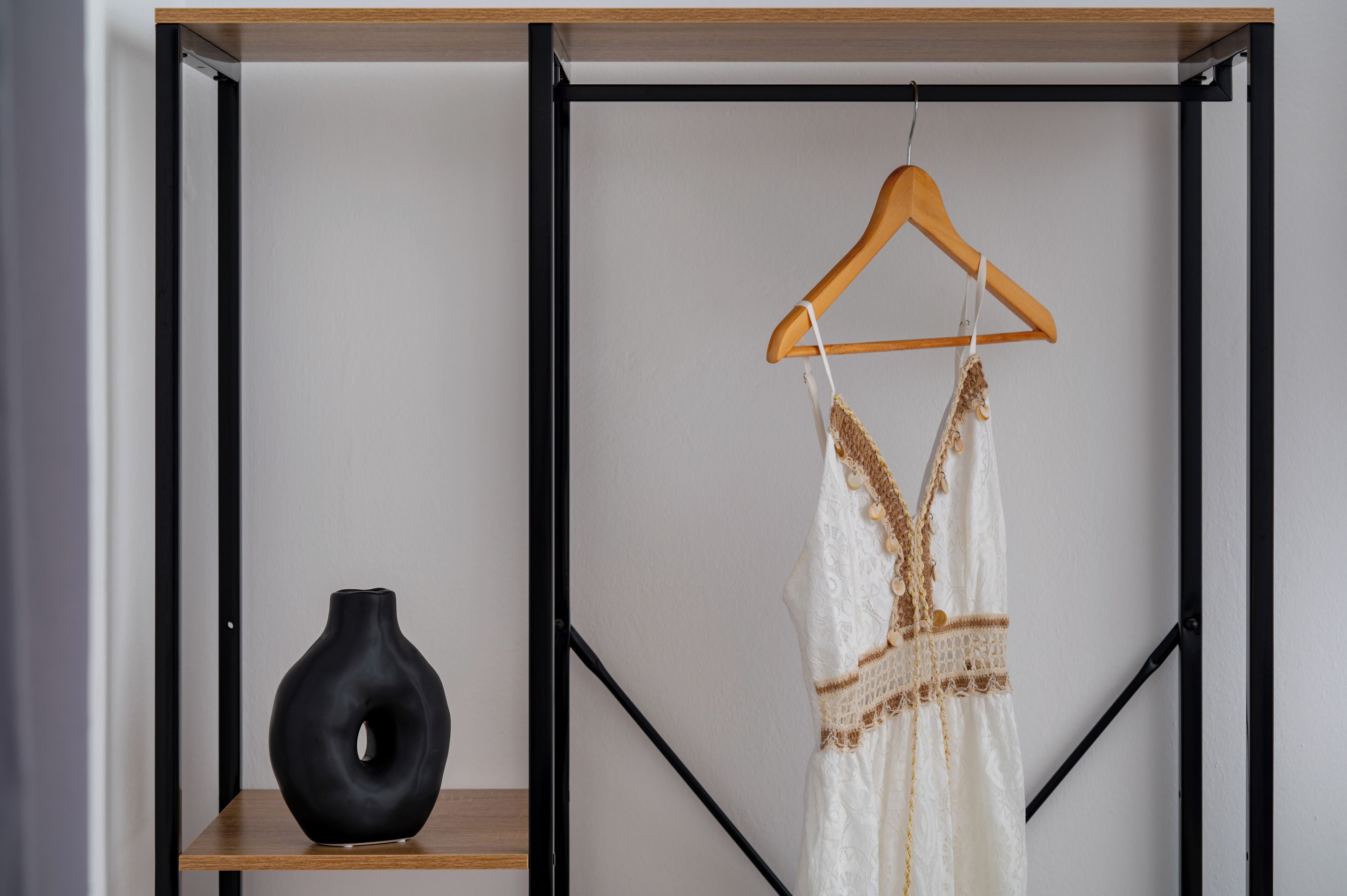 Een kamer met een zwarte vaas op een houten plank, een witte jurk hangend aan een houten kapstok, en een zwarte metalen frameconstructie.