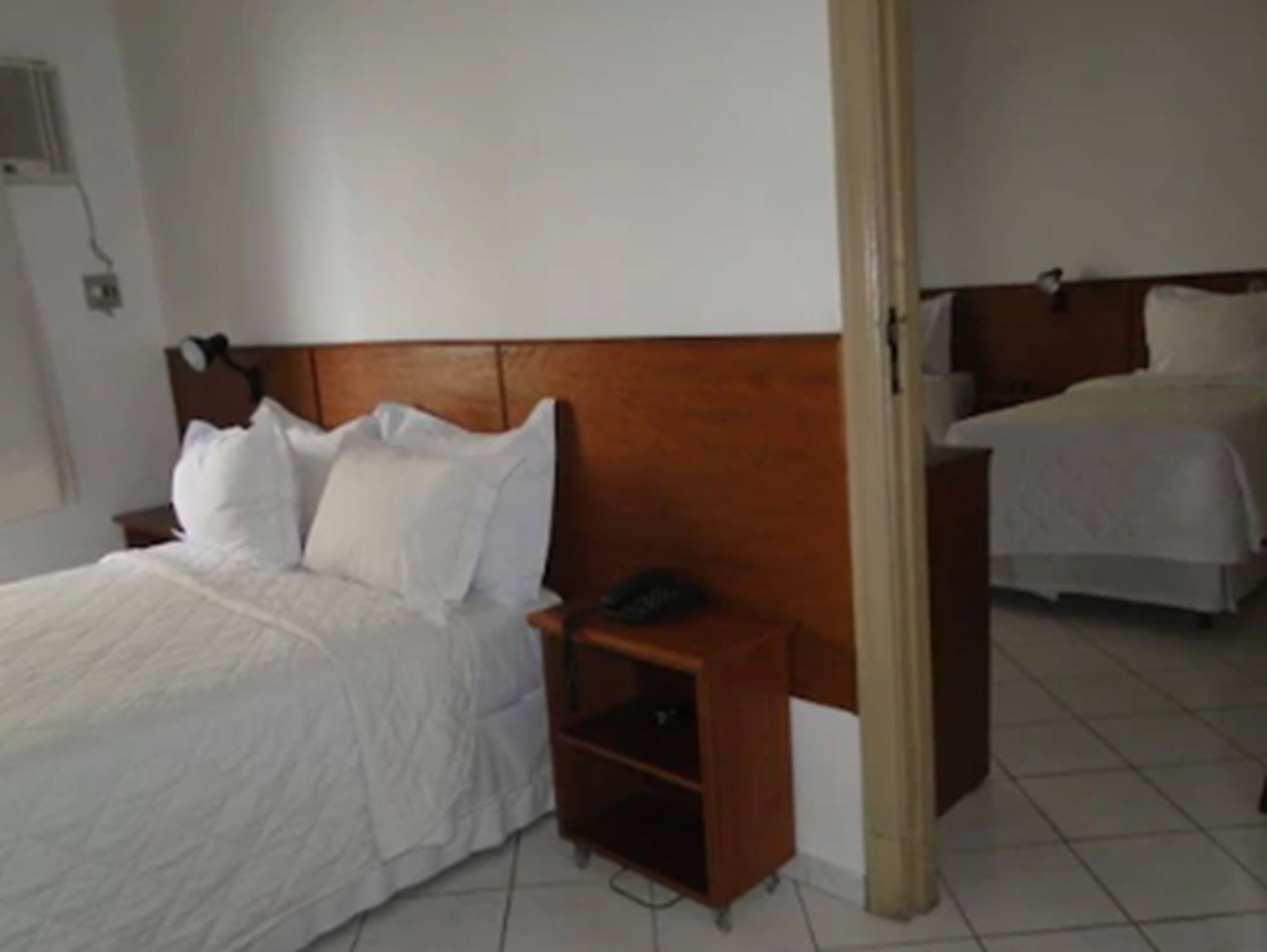 Una habitación de hotel con cabecera de madera, una cama individual con ropa de cama blanca, una mesita de noche con un teléfono y una pequeña estantería.