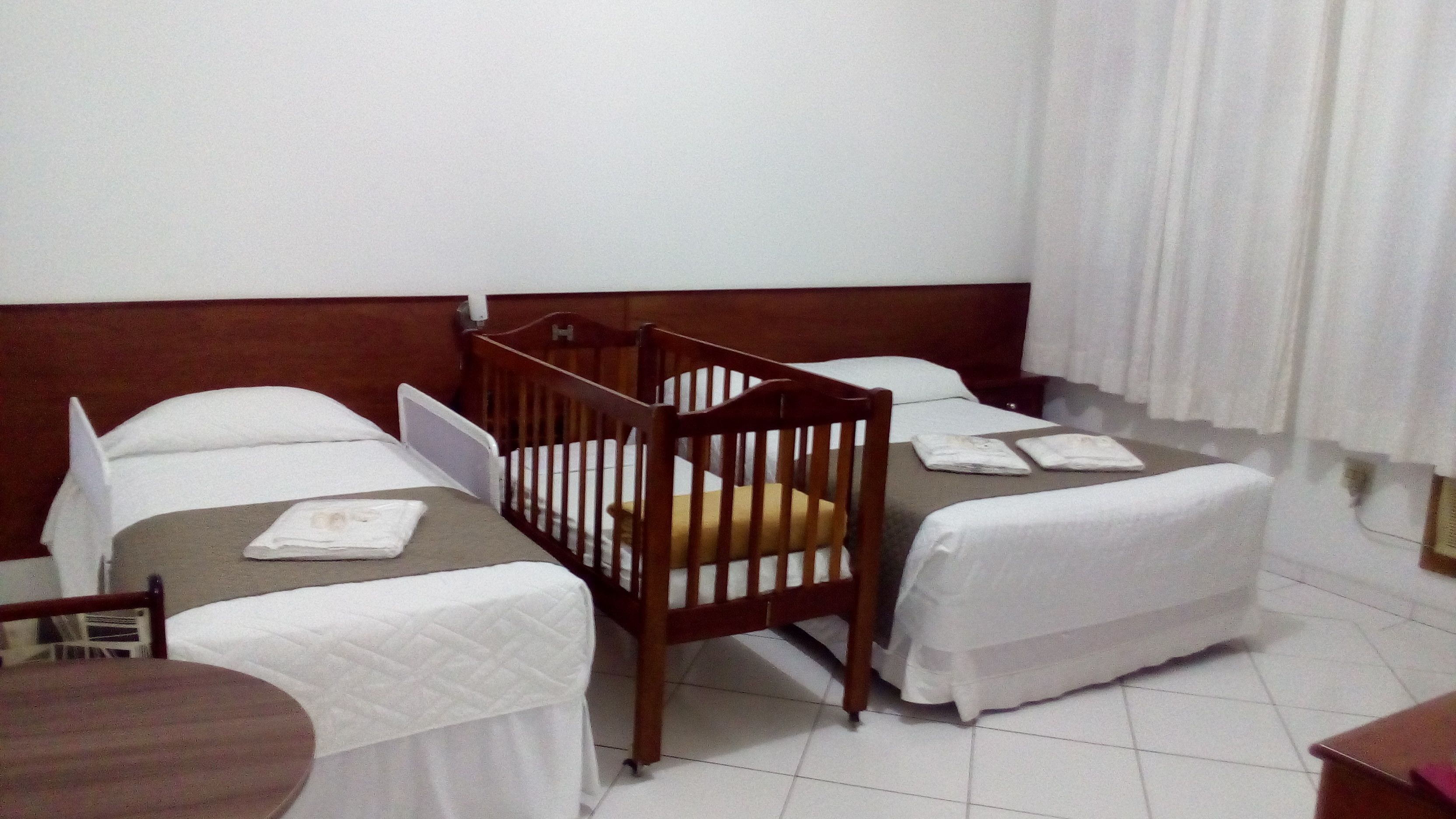 A hotel room with two single beds, a wooden crib, a small table, and a window with curtains.