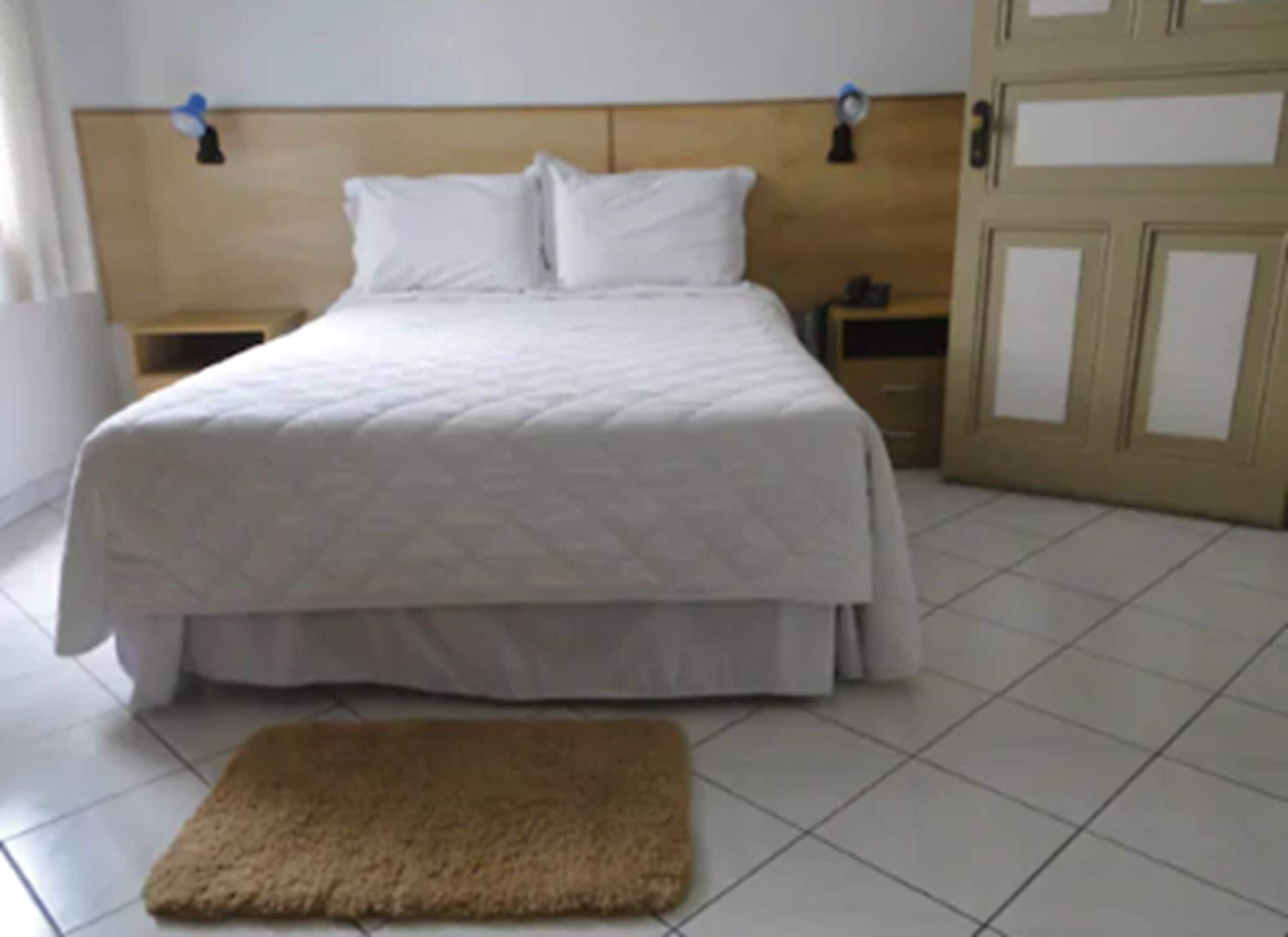 Een net opgemaakt bed met een houten hoofdbord, een klein nachtkastje en een bruin vloerkleed op een betegelde vloer.
