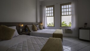 Habitación de hotel con dos camas, una mesita de noche con lámpara, una pequeña mesa con un vaso de agua y vistas a palmeras a través de las ventanas.