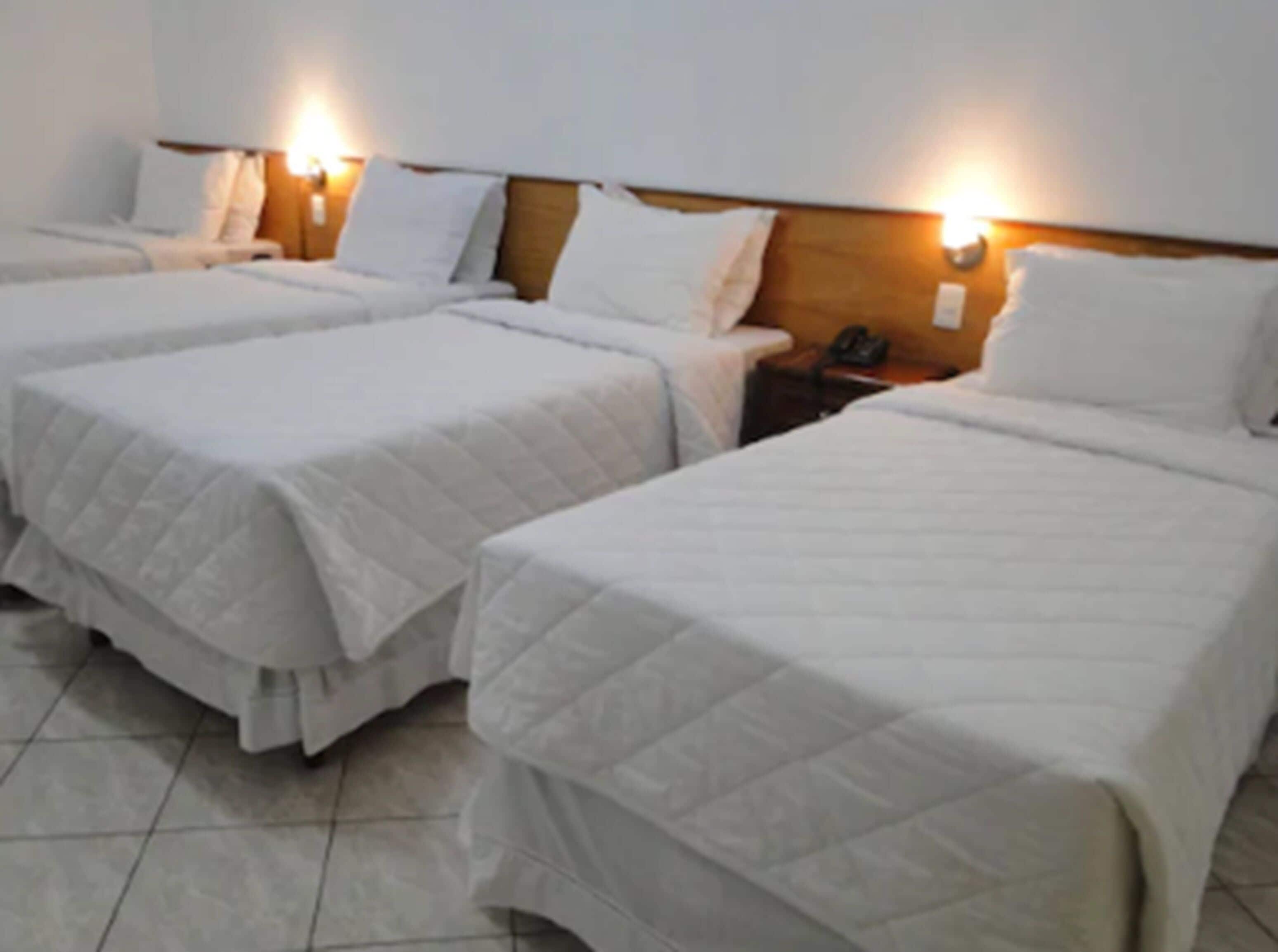 Quarto de hotel com três camas de solteiro, cada uma com colcha e travesseiros brancos. Possui cabeceira de madeira com luzes fixadas na parede sobre cada cama.