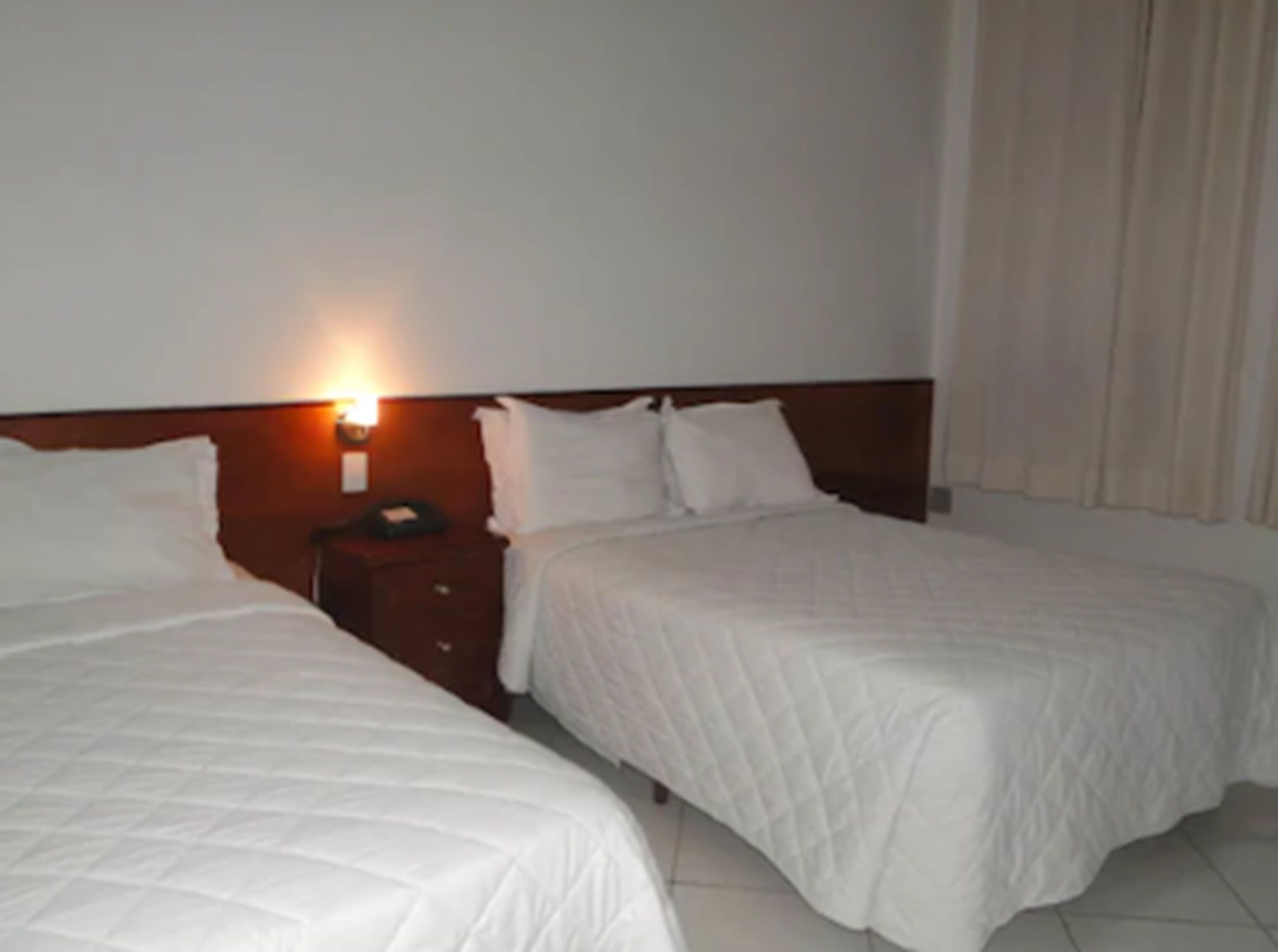 A hotel room with two beds, a nightstand, and a wall-mounted lamp.