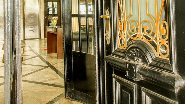 Una puerta negra ornamentada con detalles dorados intrincados, que da acceso a un vestíbulo con piso de tablones y un reloj en la pared.