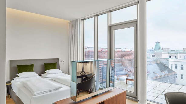 A modern hotel room with a large bed, a desk with a mirror, and a view of the city through large windows. - MAXX by Steigenberger Vienna (Vienna)