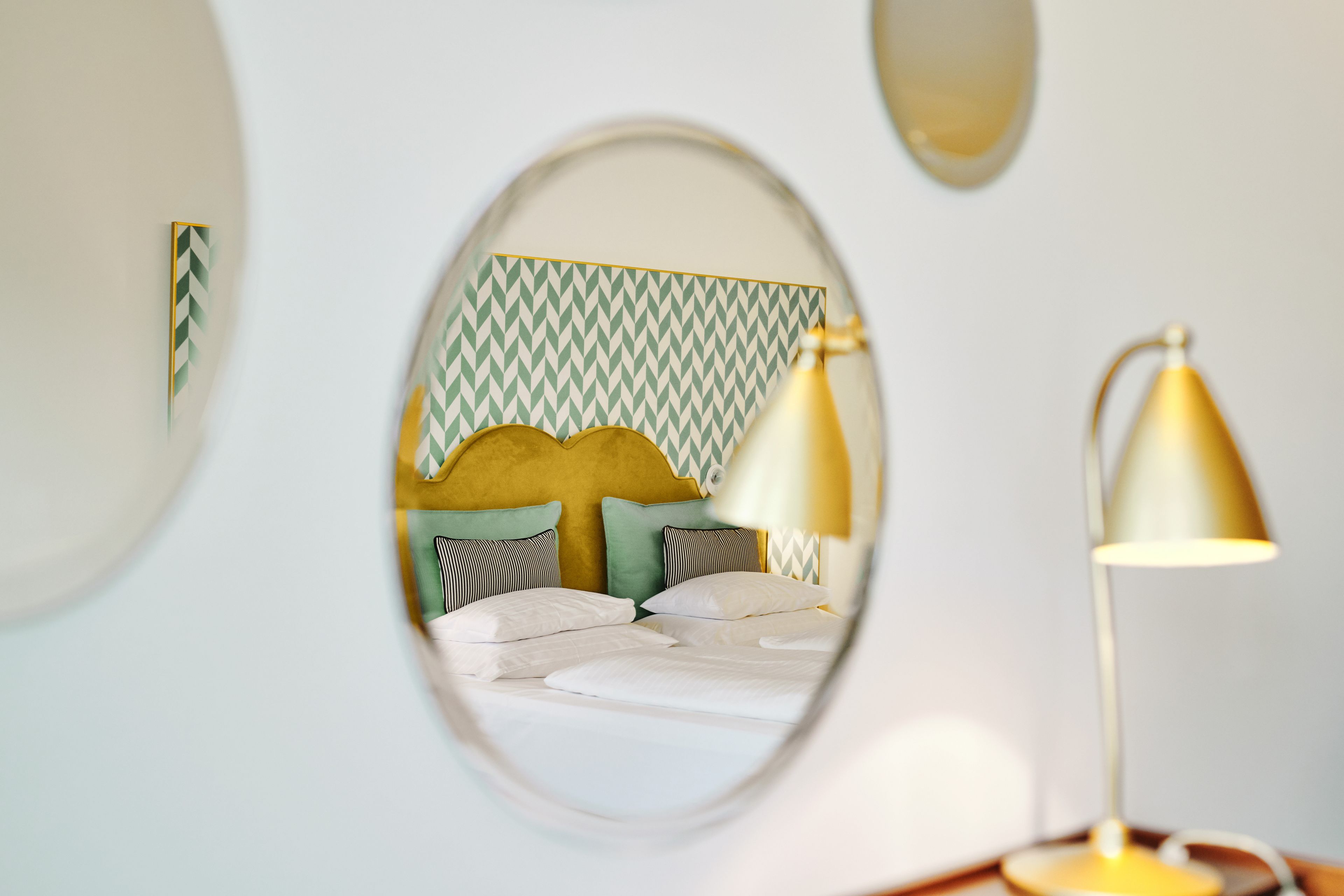 Una cama con ropa de cama blanca y un cabecero estampado, una lámpara y un espejo que refleja parte de la habitación.