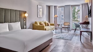 A hotel room with a large bed, a sofa, a desk, and a chair. There is a window with a view of buildings and a door leading to an outdoor area. - Crowne Plaza Vienna by IHG (Vienna)