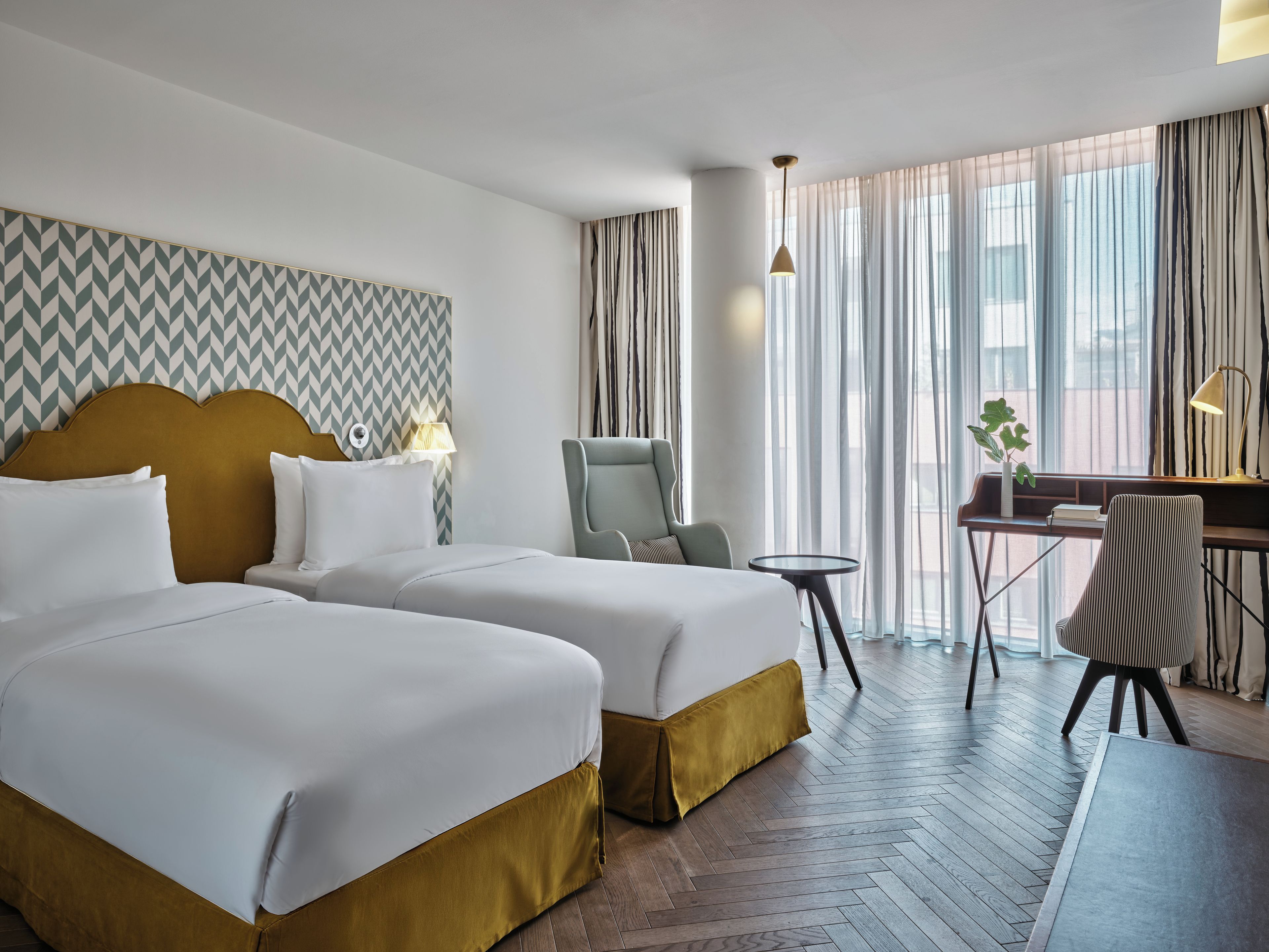 A hotel room with two beds, a desk, a chair, and a view of the city through large windows.