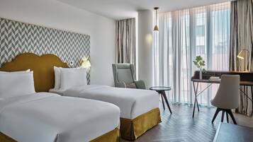 A hotel room with two beds, a desk, a chair, and a view of the city through large windows.