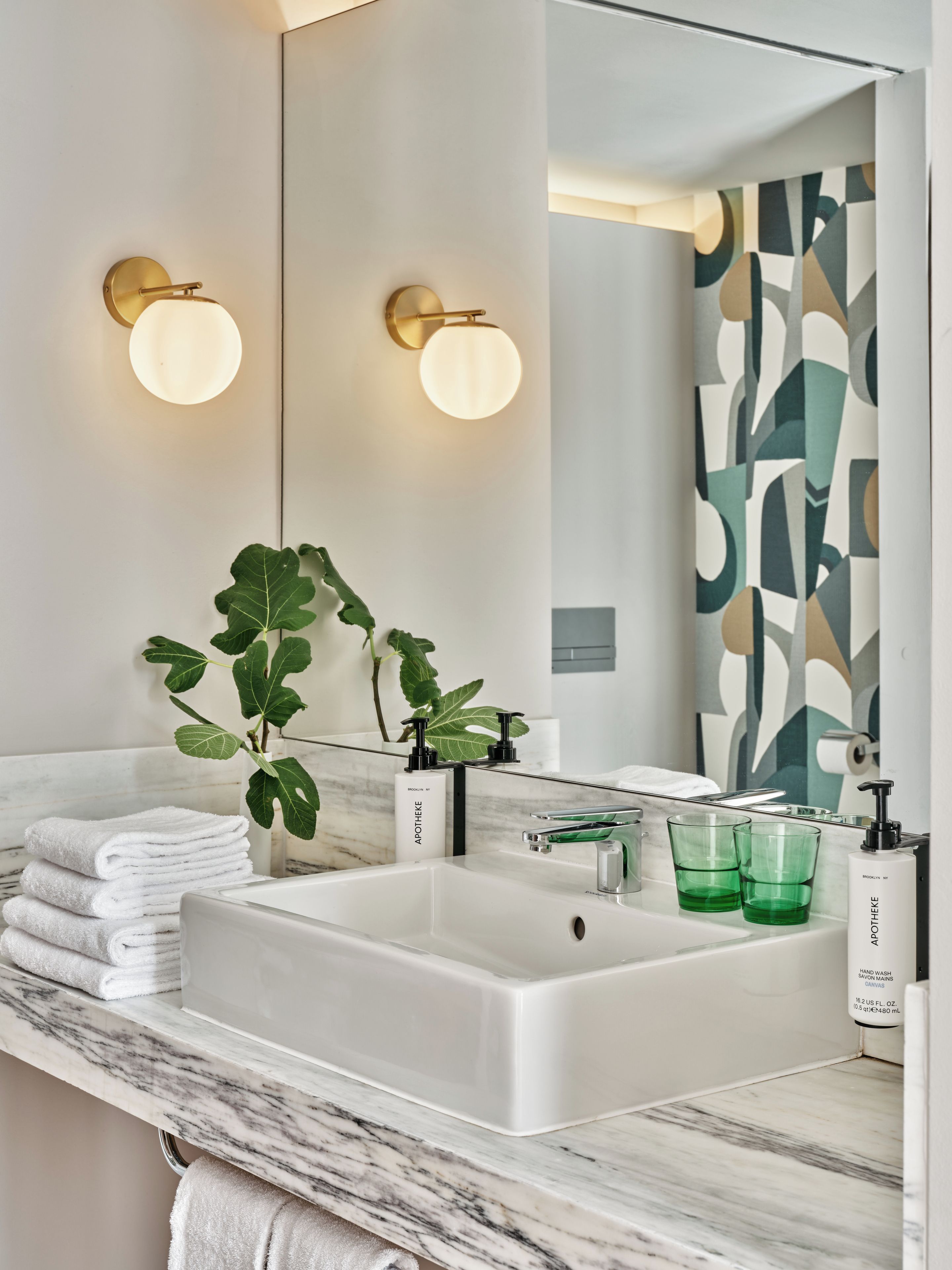Ein Badezimmer mit einem weißen Waschbecken, einer Marmorarbeitsplatte, zwei Wandleuchten, einem großen Spiegel, einer Zimmerpflanze und einem Stapel Handtüchern.