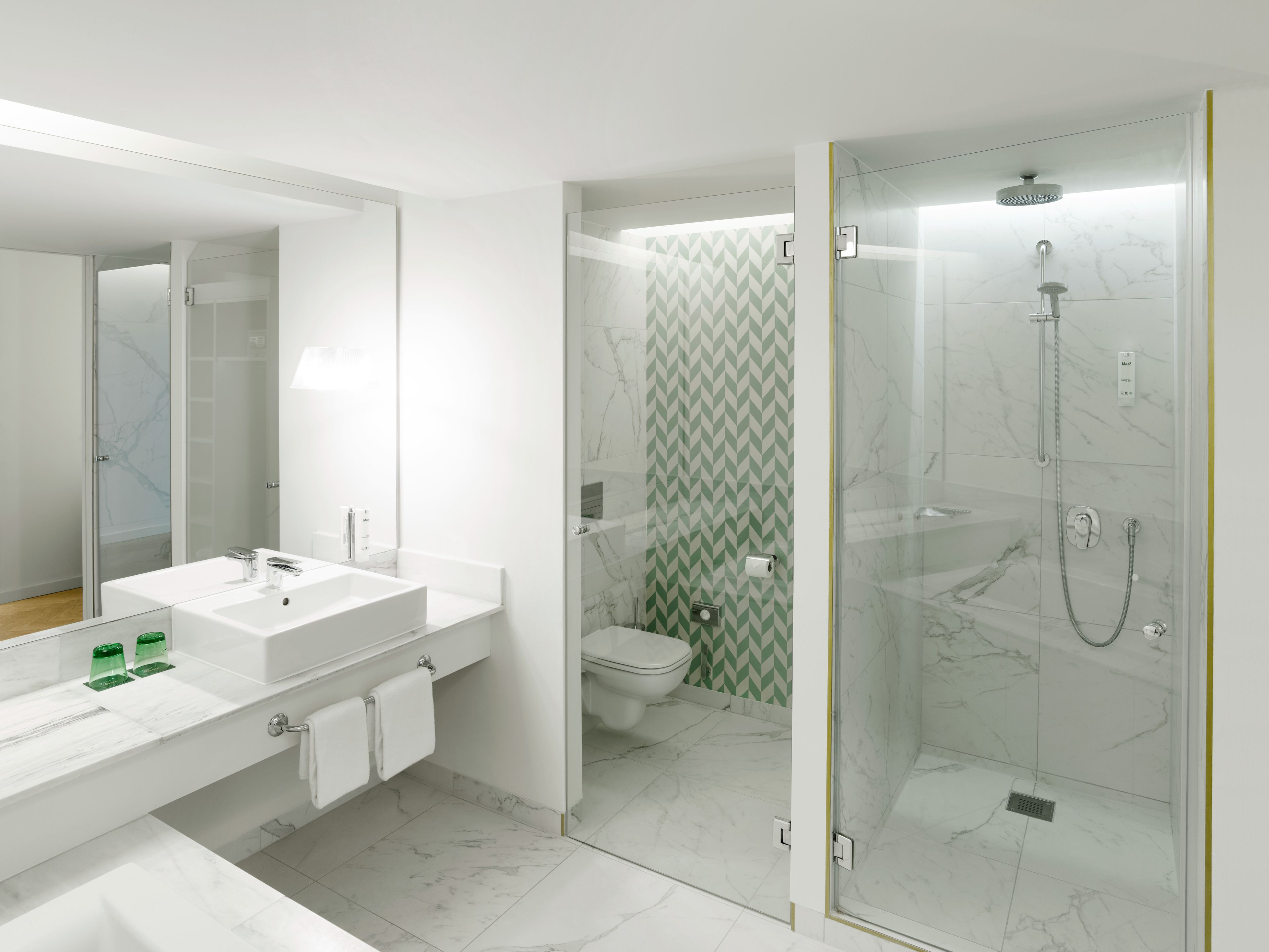 Ein modernes Badezimmer mit einer gläsernen Duschkabine, einem weißen Waschbecken mit Spiegel und einer Toilette mit grünem Wandmuster.