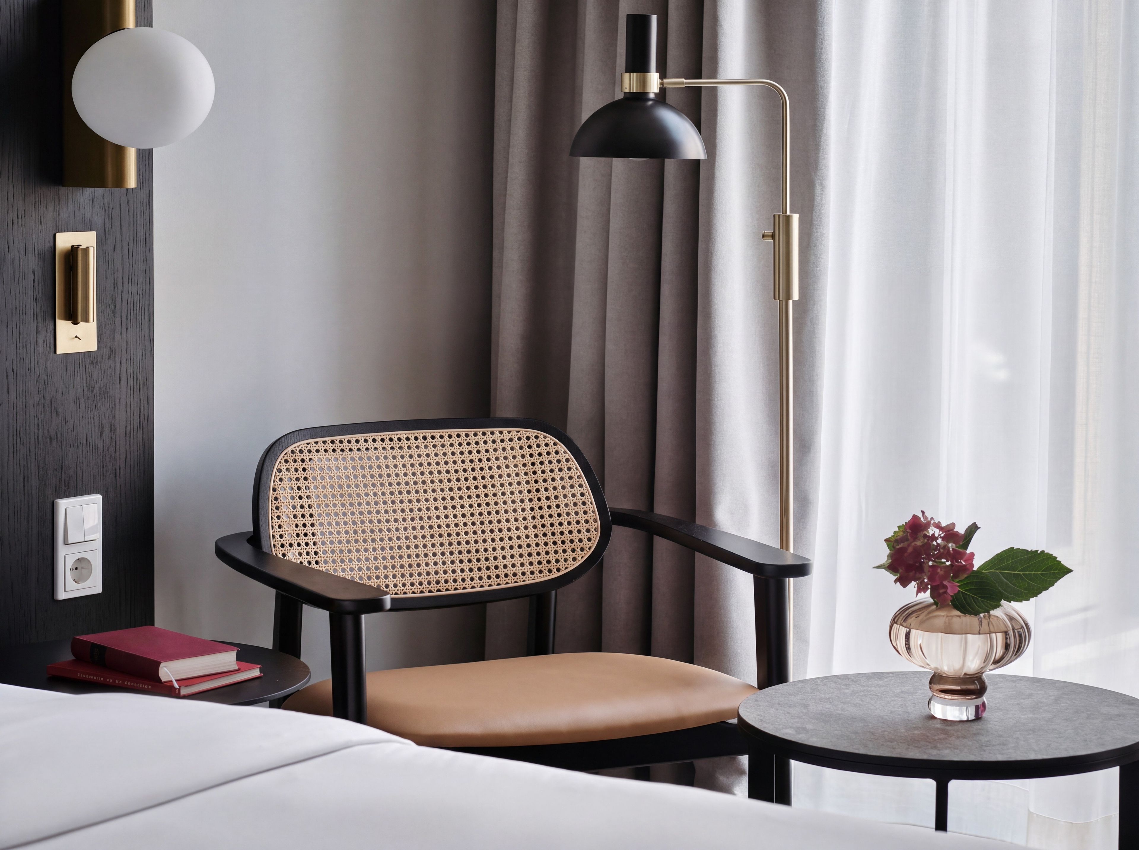 A modern hotel room with a rattan chair, a bedside table with a vase, and a floor lamp.