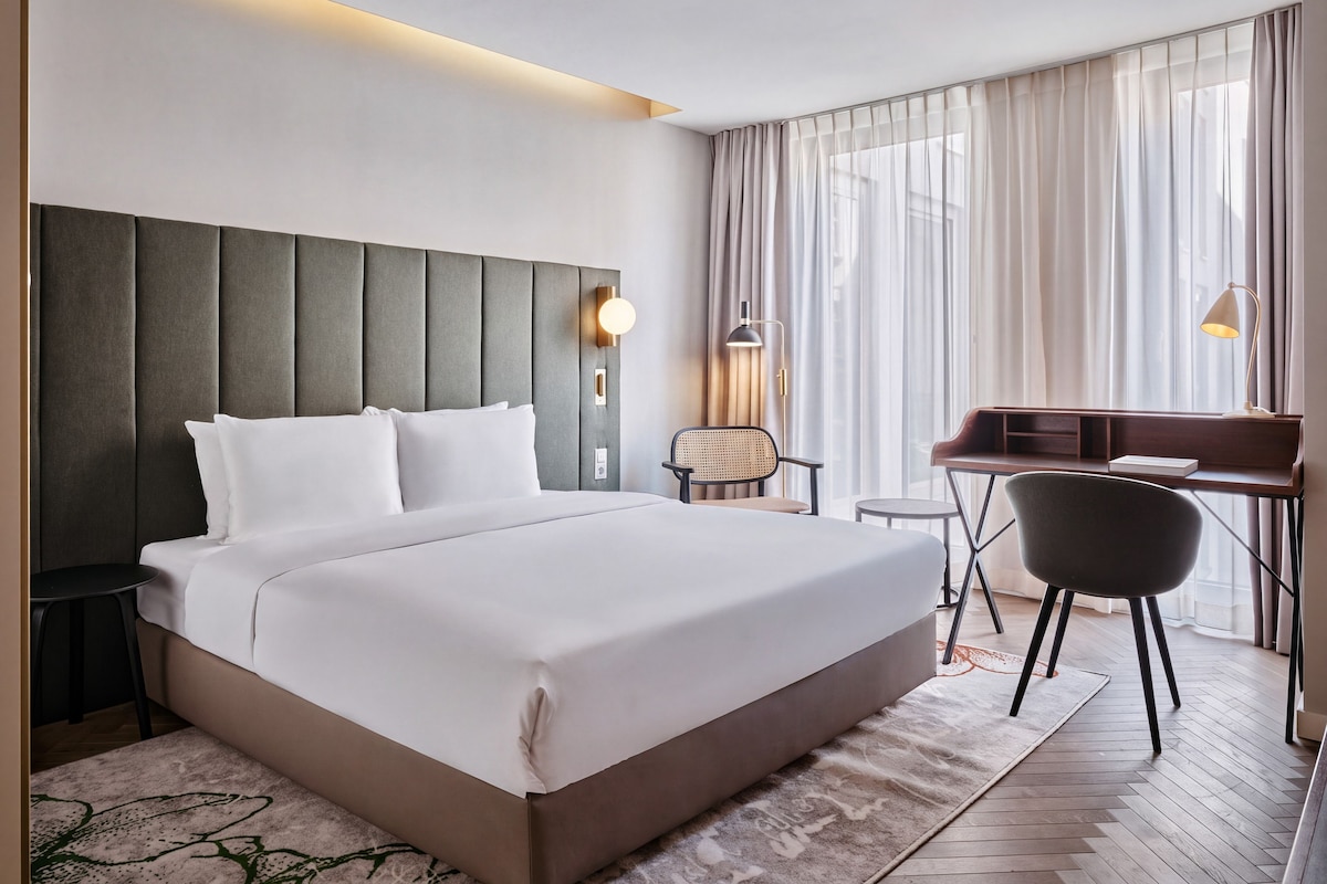 A modern hotel room with a large bed, a desk, a chair, and a view of the city through sheer curtains.