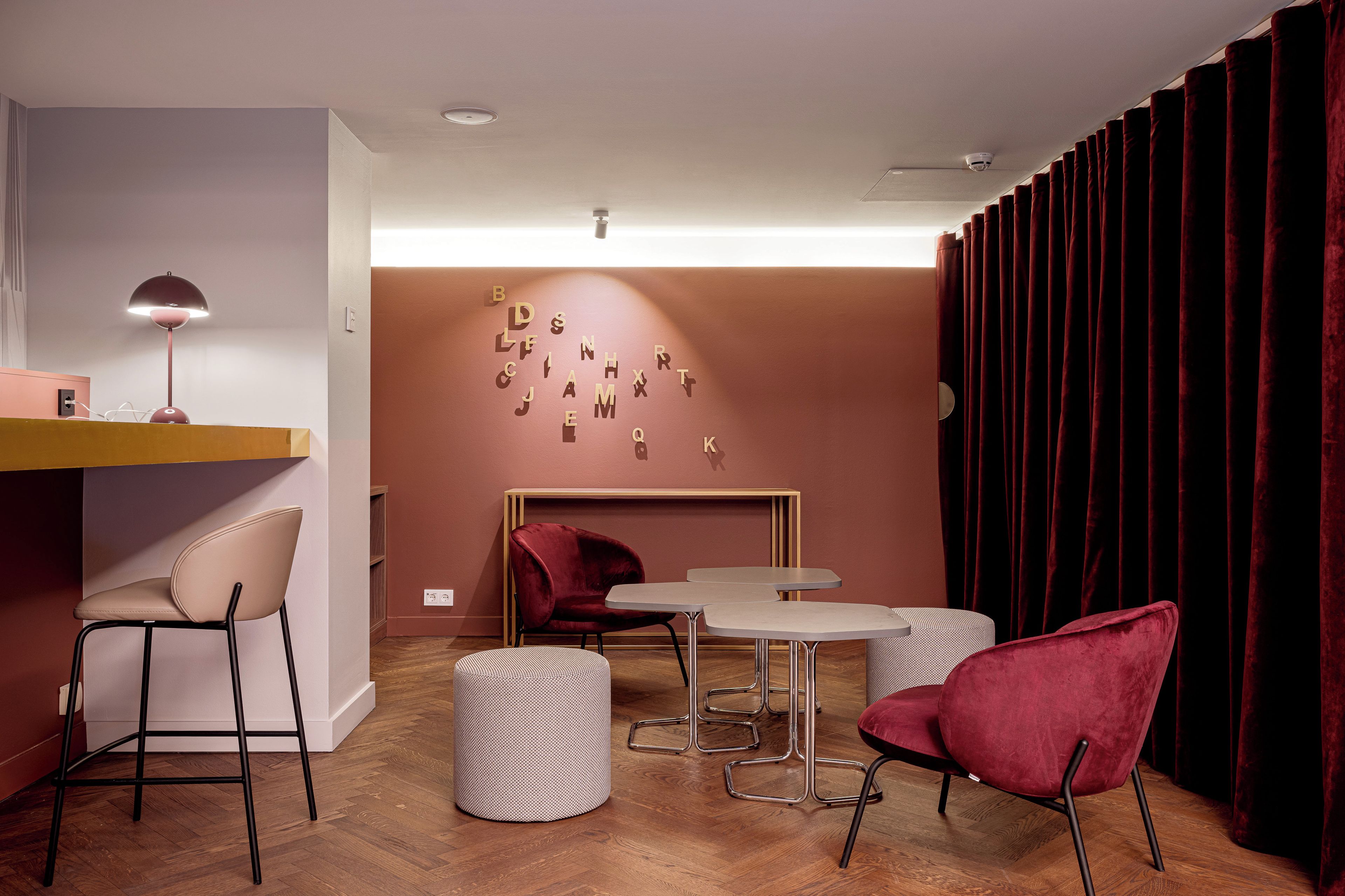 Ein modernes Zimmer mit Schreibtisch, zwei Stühlen, einem kleinen Tisch, einem roten Samtstuhl, einem gemusterten Ottomanen und einer Wand mit Buchstaben.