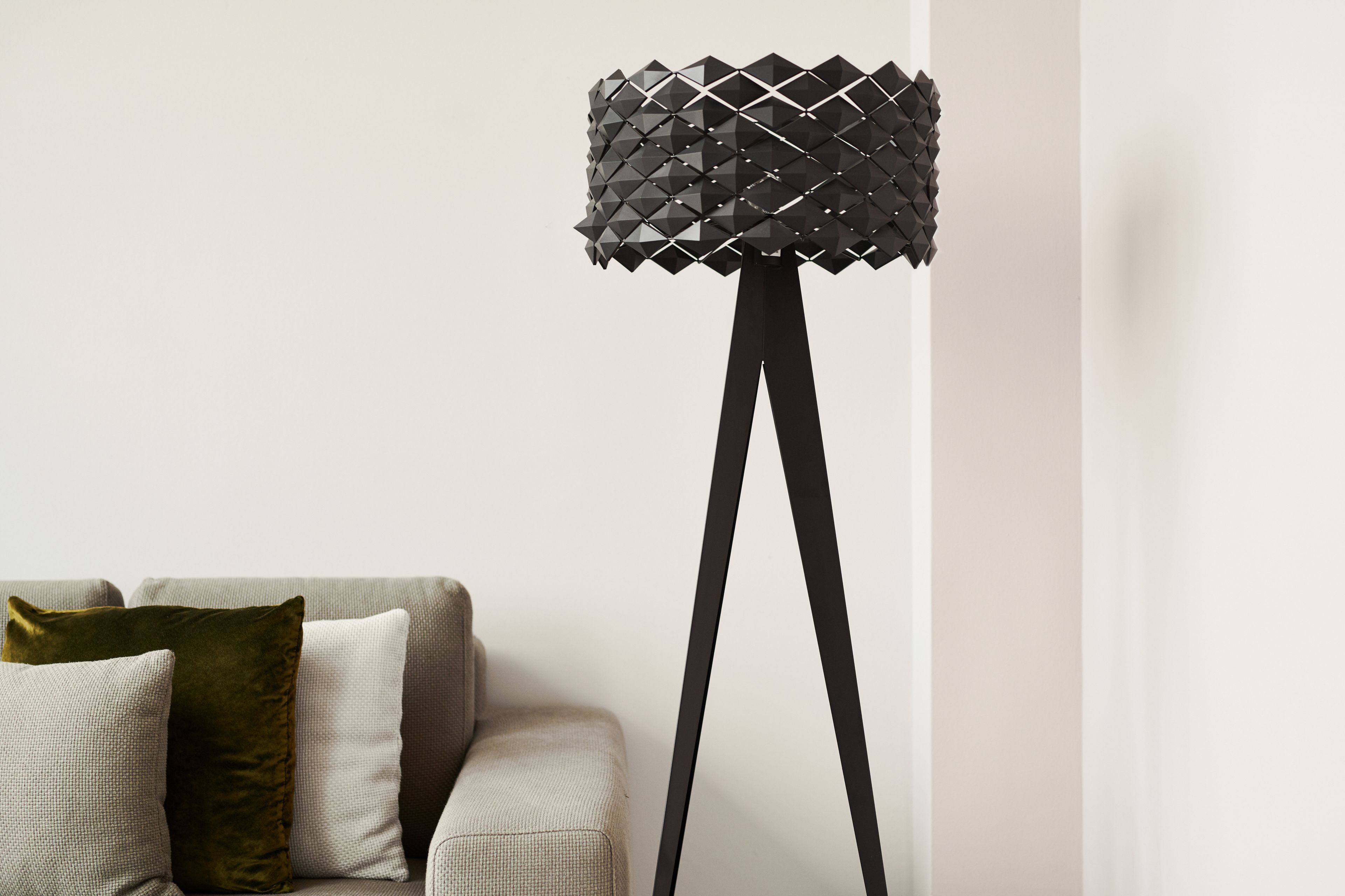 Una lampada da terra moderna con un design geometrico, un divano con cuscini decorati e una parete neutra sullo sfondo.