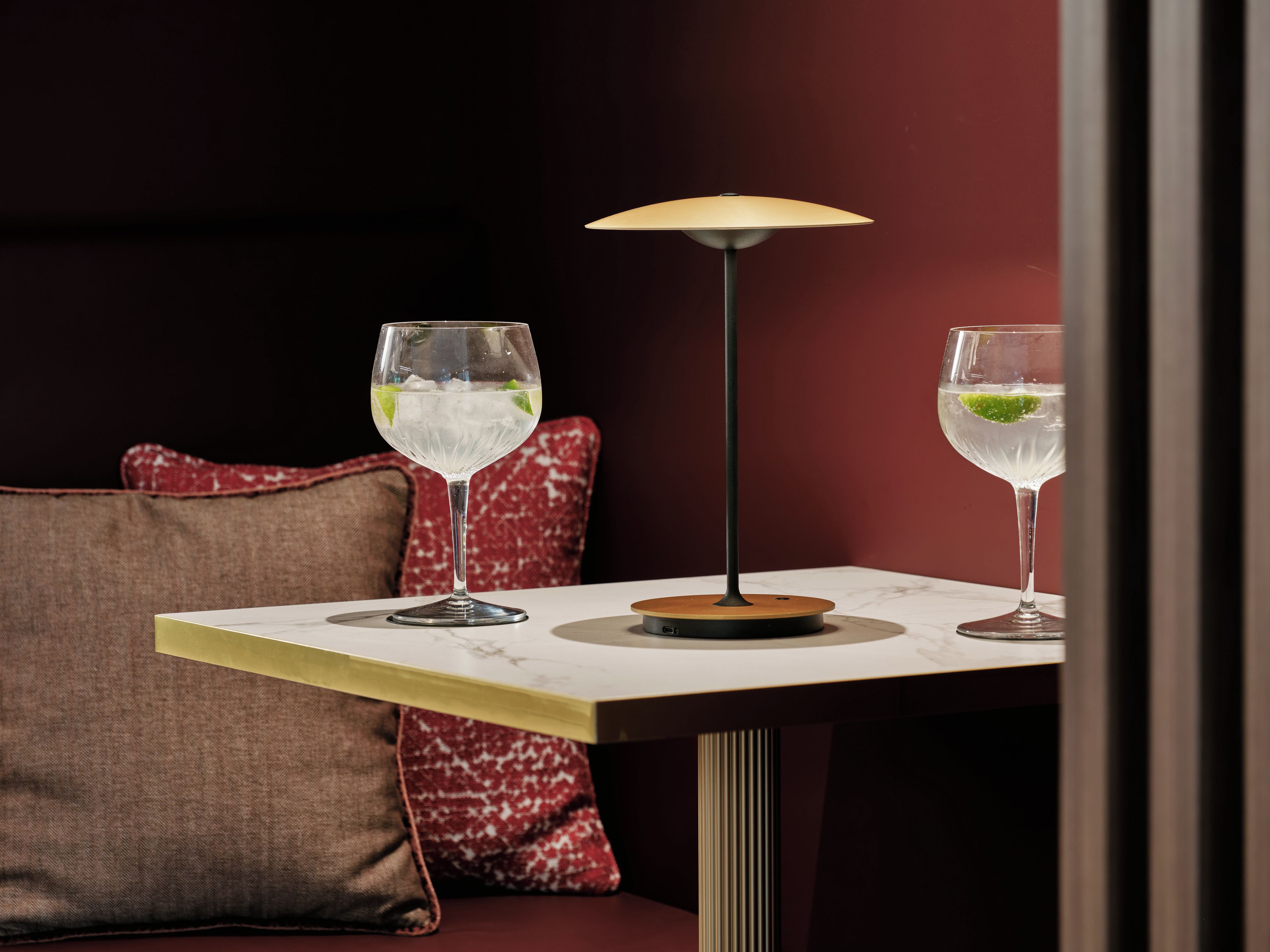 A table with two glasses of a clear beverage, a lamp, and a red upholstered bench.