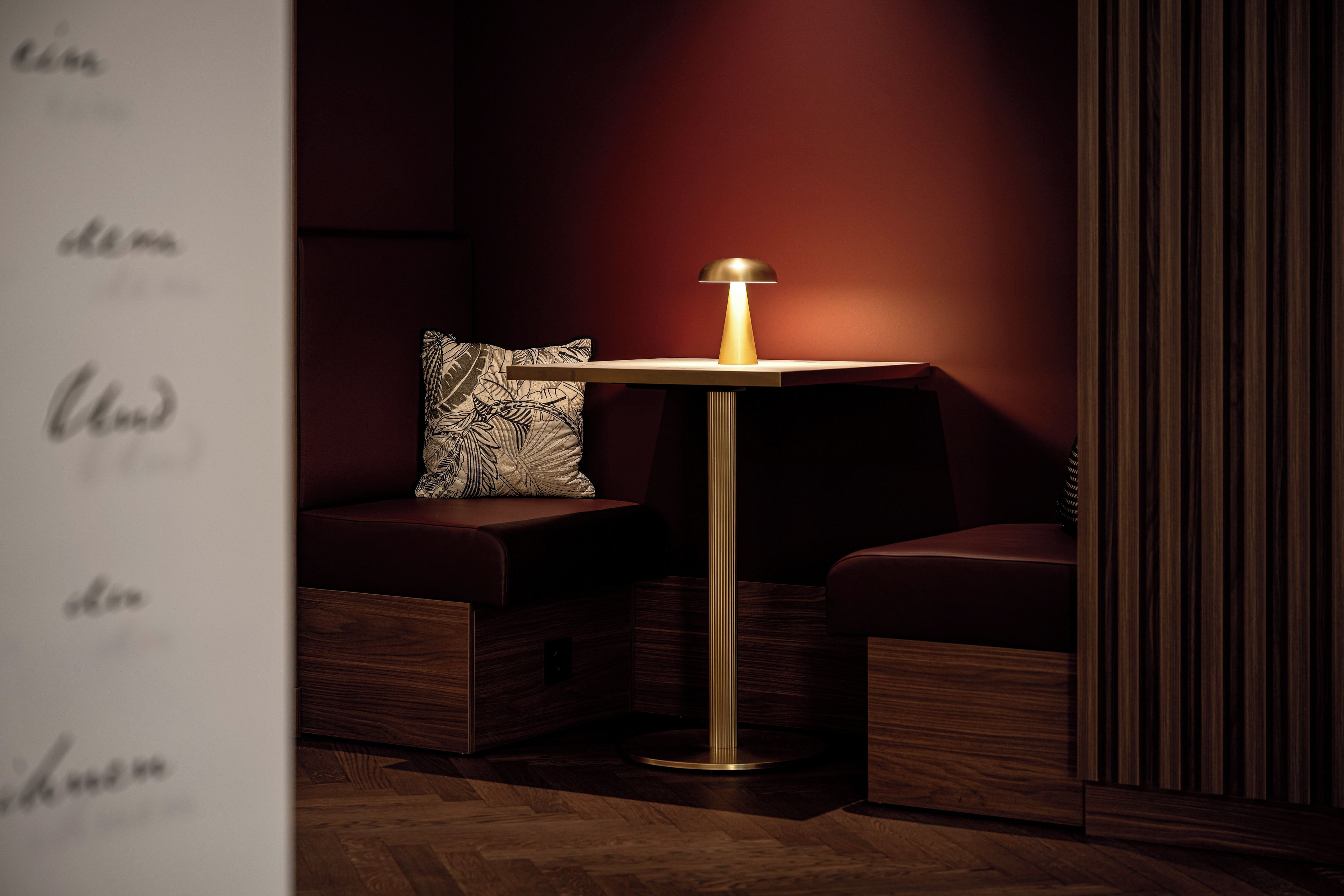 Una habitación moderna con suelo de madera, una mesita con una lámpara y un cojín estampado sobre un banco acolchado.