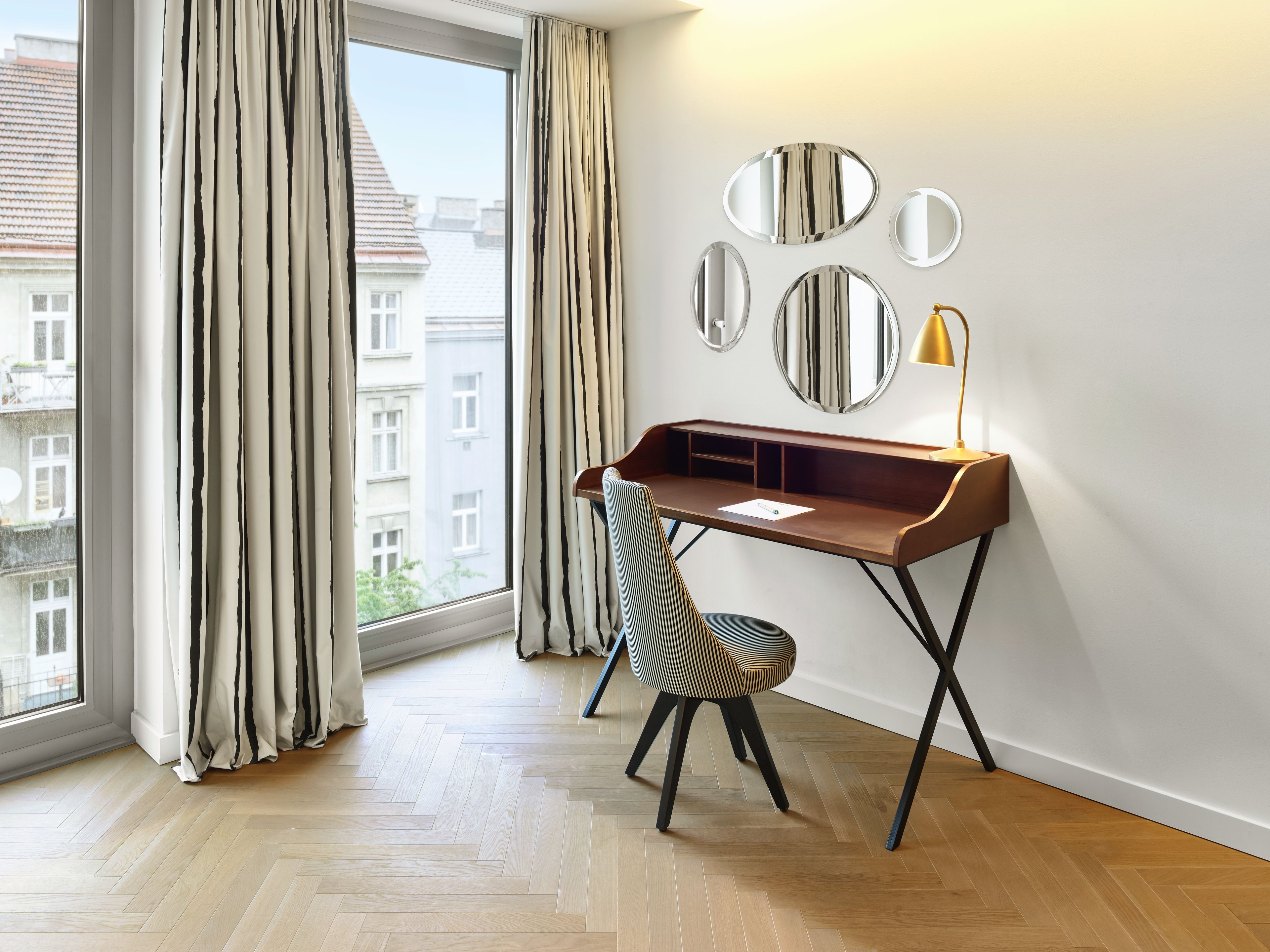 A room with a wooden desk, a chair, a lamp, and a view of buildings through a window with curtains.