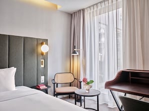 Una habitación de hotel con cama, escritorio, silla, pequeña mesa con un florero y una lámpara.