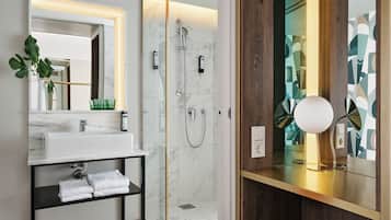 Ein modernes Badezimmer mit einer Duschkabine aus Glas, einer Marmorwand, einem weißen Waschbecken mit Ablagefläche in Schwarz und einem Holz-Waschtisch mit Spiegel.