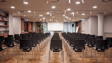 Un salón de conferencias con filas de sillas, una pantalla de proyector y un escenario.