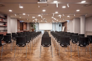 Uma sala de conferências com fileiras de cadeiras, uma tela de projetor e um palco.
