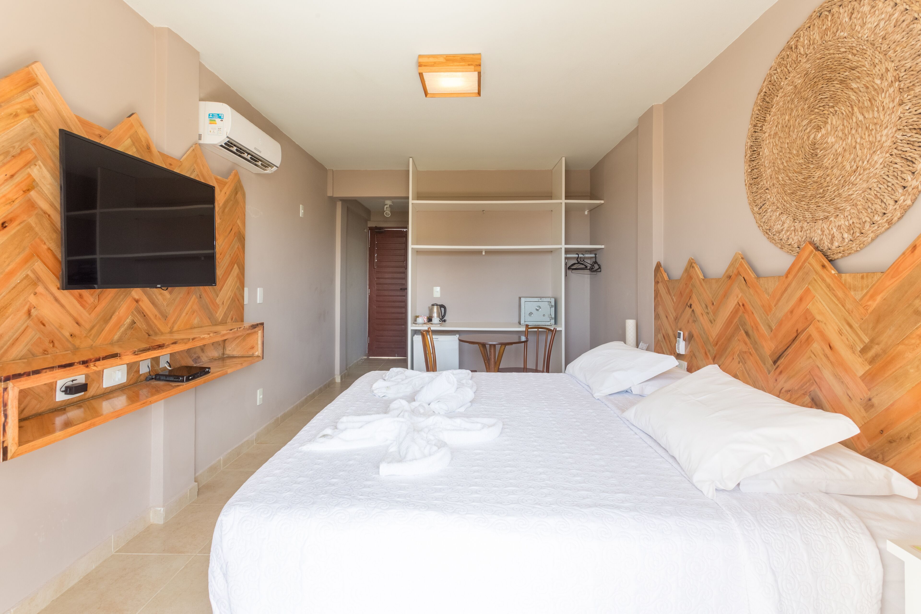 A hotel room with a bed, a TV mounted on a wooden wall, a desk with a chair, and a wardrobe.