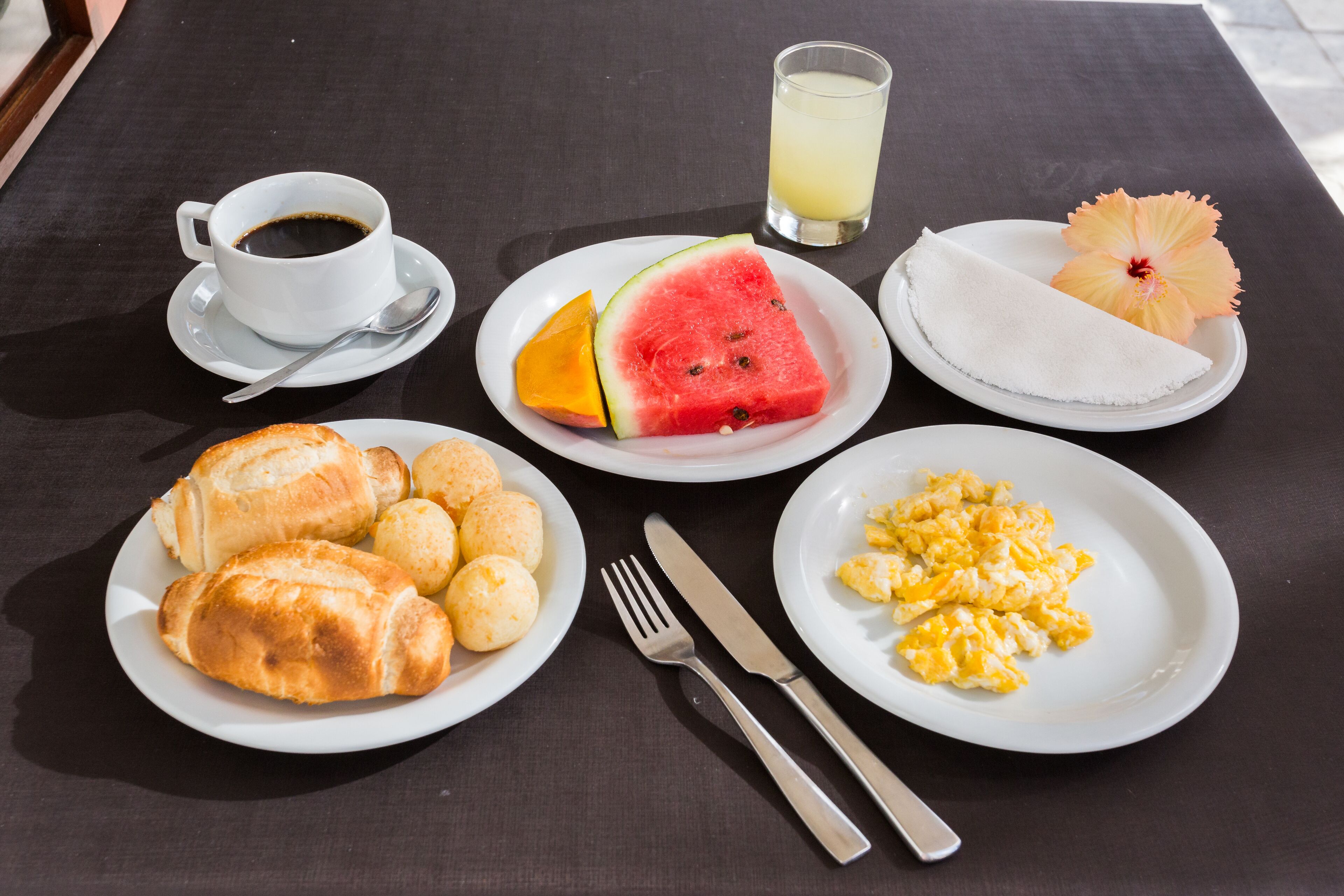 Una mesa de desayuno con platos de cruasanes, huevos revueltos, sandía, naranja y un vaso de jugo.